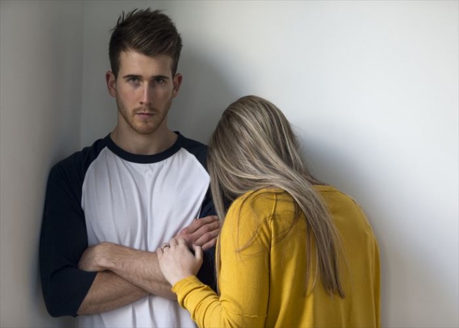 Young woman crying on her partners shoulder. He is looking at the camera with his arms folded.