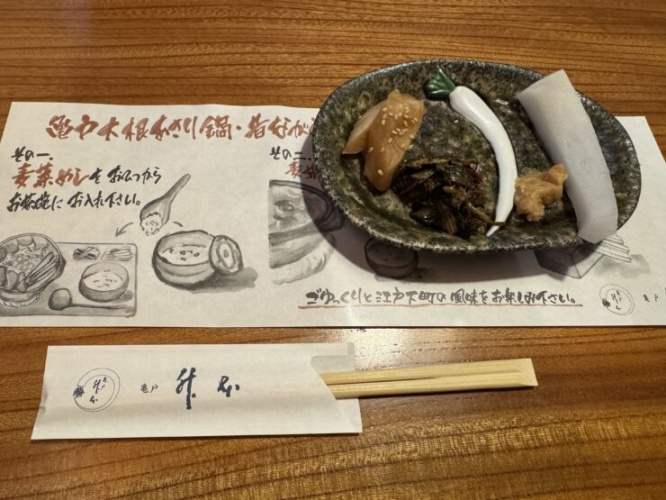 Ceramic plate with pickled vegetables topped with sesame seeds and a white fish cake strip, accompanied by chopsticks and printed menu sheets at a dining table.