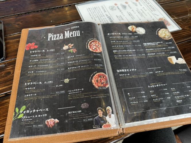 Open pizza menu on a dark board, showing pizza images, Japanese text, and prices on a wooden table.