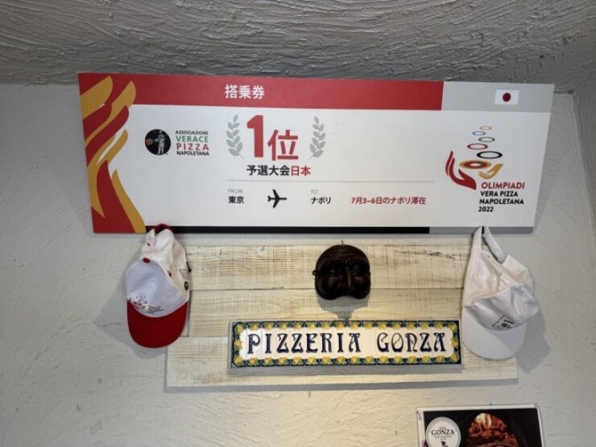 A wall-mounted signboard for a Neapolitan pizza restaurant with logos, Japanese text, and colorful decorative elements, flanked by a red cap and a white apron hanging nearby.