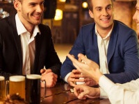 Three young businessmen in suits are smiling, talking and drinking beer while sitting in pub