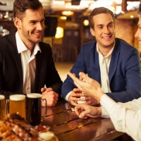 Three young businessmen in suits are smiling, talking and drinking beer while sitting in pub