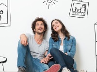 Portrait Of Happy Young Couple Sitting On Floor Looking Up While Dreaming Their New Home And Furnishing