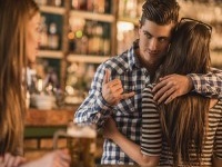 Unfaithful young man in a cafe. He is embracing his girlfriend and making call gesture to another woman behind her back.