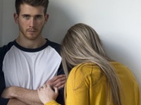 Young woman crying on her partners shoulder. He is looking at the camera with his arms folded.