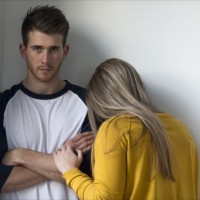 Young woman crying on her partners shoulder. He is looking at the camera with his arms folded.
