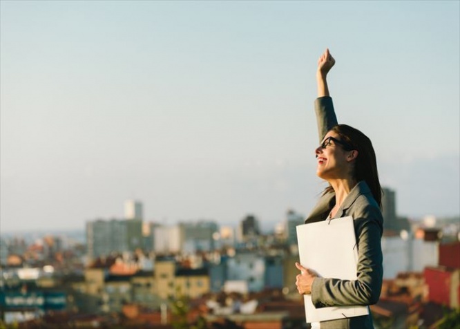 Successful young businesswoman raising arm celebrating business  or job achievement towards city background. Professional happy woman walking outside.