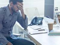 Stressed business man at the office. He is casually dressed and looking distraught. He looks very uncomfortable and could also have a headache. He is has his head in his hand and looks very upset. Hi is sitting at a desk with a computer and phone. Copy sp