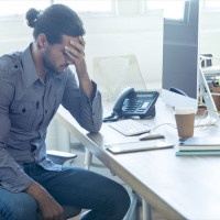 Stressed business man at the office. He is casually dressed and looking distraught. He looks very uncomfortable and could also have a headache. He is has his head in his hand and looks very upset. Hi is sitting at a desk with a computer and phone. Copy sp