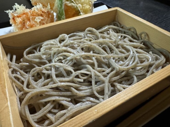 Soba noodles in a wooden box with assorted tempura pieces behind them.