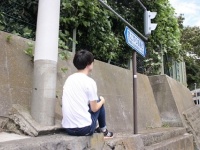 尻で押すほど重い物を持っていなかったので、標識の下で体育座りをして、尻で坂を体感してみた記者。三脚とタイマー機能による撮影後、坂を上ってきた歩行者に訝しげに一瞥された（神奈川県横須賀市にて。以下記者撮影）