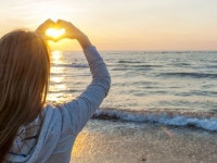 Blonde young girl holding hands in heart shape framing setting sun at sunset on ocean beach