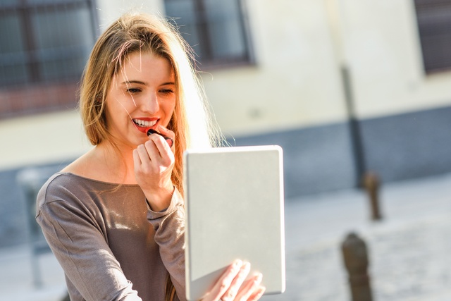 Young woman applying lipstick looking at tablet