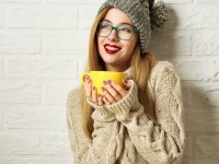 Happy Smiling Hipster Girl in Knitted Sweater and Beanie Hat with a Mug in Hands. Beautiful Woman at White Brick Wall Background. Winter or Autumn Warming Up Concept. Toned Photo with Copy Space.