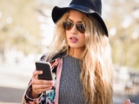 Young woman looking at her smartphone in urban background. Girl wearing jacket, hat, sweater and sunglasses.