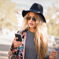 Young woman looking at her smartphone in urban background. Girl wearing jacket, hat, sweater and sunglasses.