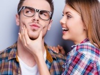 Handsome young man grimacing while his girlfriend touching his face while standing against grey background