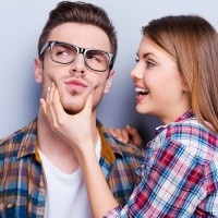 Handsome young man grimacing while his girlfriend touching his face while standing against grey background