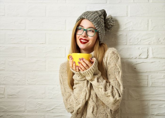 Happy Smiling Hipster Girl in Knitted Sweater and Beanie Hat with a Mug in Hands. Beautiful Woman at White Brick Wall Background. Winter or Autumn Warming Up Concept. Toned Photo with Copy Space.