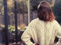 A young woman is standing by the french doors in her house and is looking out at the rain