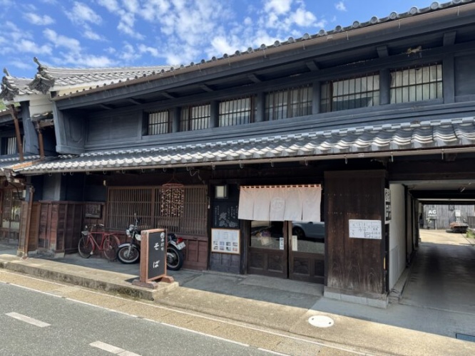 Traditional Japanese shopfront with dark wooden exterior, tiled roof, and white fabric noren curtains outside the entrance. Bicycles parked nearby.