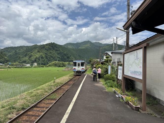 Two people stand on a rural train platform beside tracks, with a small blue-and-white train and green hills in the distance under a cloudy sky.