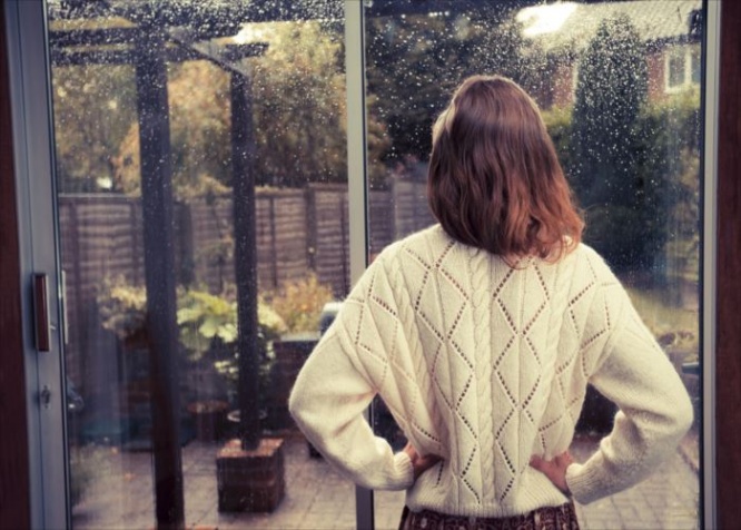 A young woman is standing by the french doors in her house and is looking out at the rain