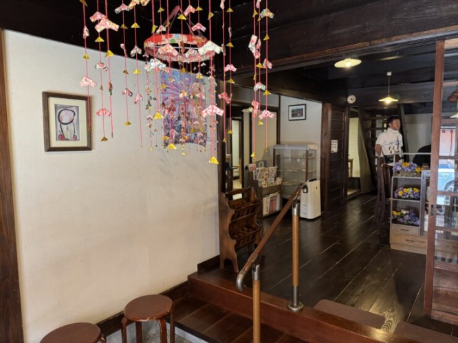 Interior of a cozy cafe with colorful hanging origami decorations and string lights, a wooden staircase and shelves in the background, and a person in white clothing standing near the far wall.