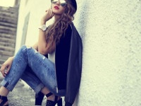 fashion model with long curly hair wearing sunglasses sitting and posing outdoor. Jeans, shoes, hat, jacket.