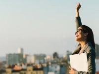Successful young businesswoman raising arm celebrating business  or job achievement towards city background. Professional happy woman walking outside.