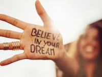 Closeup of a teenage girl's hand with the phrase "Believe in your Dreams" written in permanent marker on it, with her smiling face blurred in the background