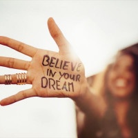 Closeup of a teenage girl's hand with the phrase "Believe in your Dreams" written in permanent marker on it, with her smiling face blurred in the background