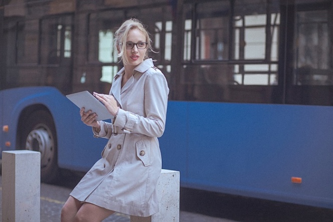 Young woman is using a digital tablet in the urban environment. She is leaning against a street pillar, while a blurred city bus passes in the background. The woman has a blonde hair that is swept back from her face and wears a beige trench coat and black eyeglasses. She smiles while looking away over the shoulder. Shallow DOF; Soft focused; developed from RAW; retouched with special care and attention; small amount of grain added for best final impression; ready made for print and web use.