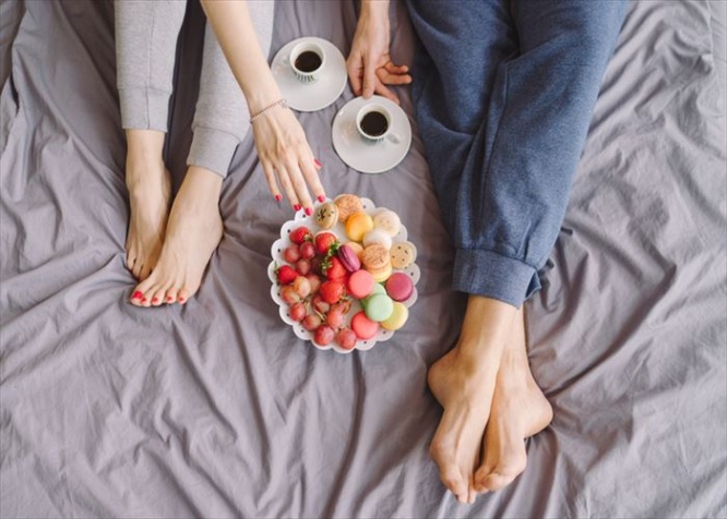 Young caucasian couple having romantic breakfast in bed. Closeup of female and male feet, two cups of coffee, fruits and colorful biscuits.
