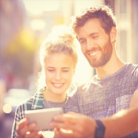 Smiling couple taking selfie through smart phone. Male and female partners spending leisure time in city. They are wearing casuals on sunny day.