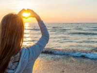 Blonde young girl holding hands in heart shape framing setting sun at sunset on ocean beach