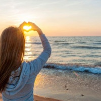 Blonde young girl holding hands in heart shape framing setting sun at sunset on ocean beach
