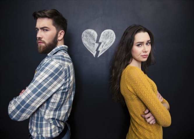Unhappy young couple standing with arms crossed over background of chalkboard with drawn broken heart