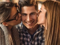 Three young people enjoying in a cafe.  Young women are kissing in cheek a man who is embracing them and looking at camera.
