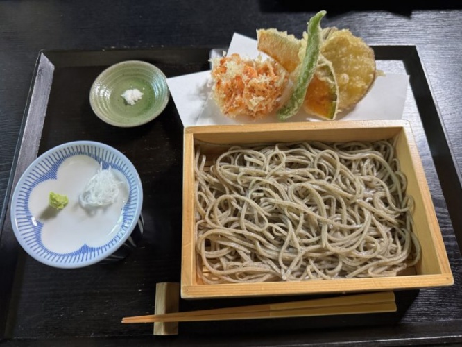 Soba noodles in a wooden box with vegetable tempura, served with wasabi and shredded radish on the side, on a dark table.