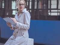 Young woman is using a digital tablet in the urban environment. She is leaning against a street pillar, while a blurred city bus passes in the background.  The woman has a blonde hair that is swept back from her face and wears a beige trench coat and blac