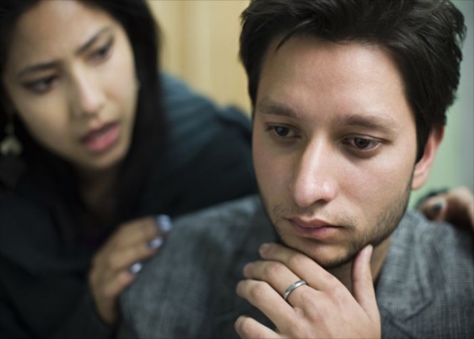 Indoor image of sad young man consoled by a young woman. Selective focus is on young man and young woman is defocused in the background, she is holding his shoulders from behind and supporting him. Two people, horizontal composition with selective focus.