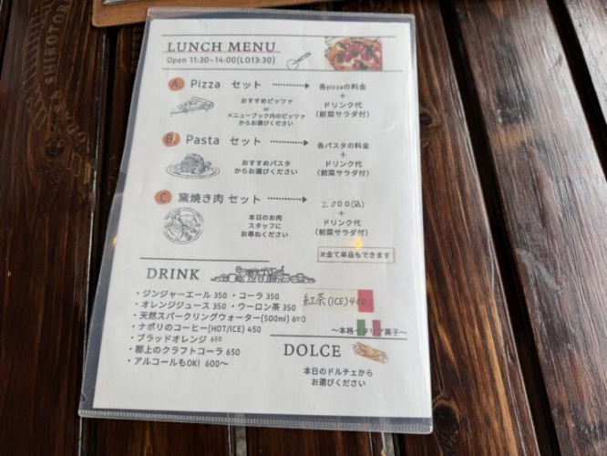 Lunch menu in a plastic sleeve on a dark wooden table, featuring pizza and pasta sections with Japanese text and small illustrations