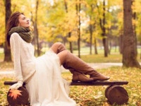 Beautiful girl sitting on a bench in park and enjoy while watching autumn colors all around. He's wearing a white skirt, black leather boots, handmade sweater and green scarf. She is relaxed, happy and looks into the distance.