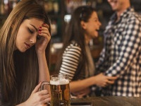 Young depressed woman sitting in the bar and holding her head in pain while happy couple is laughing in the background.