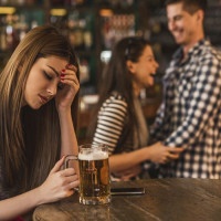 Young depressed woman sitting in the bar and holding her head in pain while happy couple is laughing in the background.