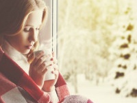 Pensive sad girl with a warming drink looking out the window in the winter forest