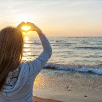 Blonde young girl holding hands in heart shape framing setting sun at sunset on ocean beach