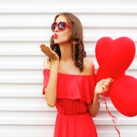 Portrait woman in red dress sends air kiss with balloon heart shape over white background