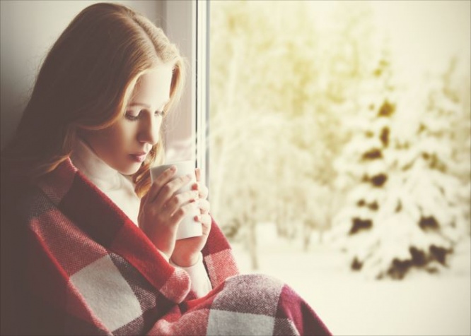 Pensive sad girl with a warming drink looking out the window in the winter forest